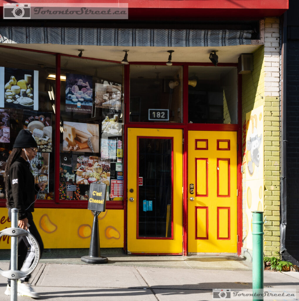 Bright yellow and red storefront of C'est Cheese shop, pedestrian walking past, bold colour in Toronto Leica Q2