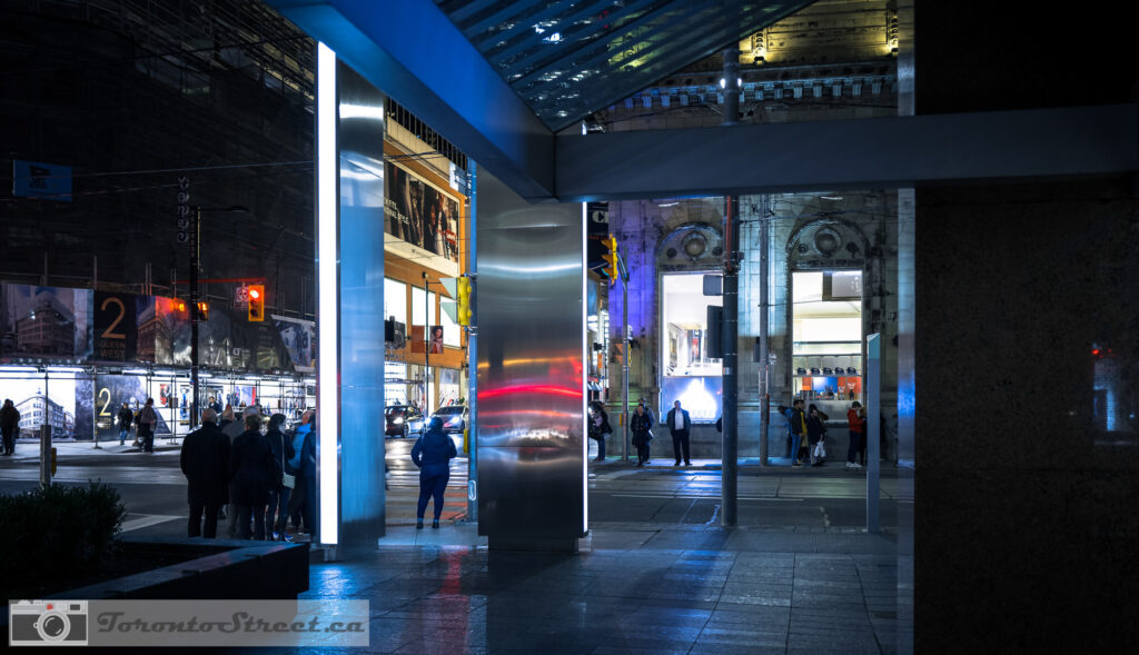 Blue-lit steel pillars near Eaton Centre at night, people silhouetted in shadow, moody colourful Toronto street photography