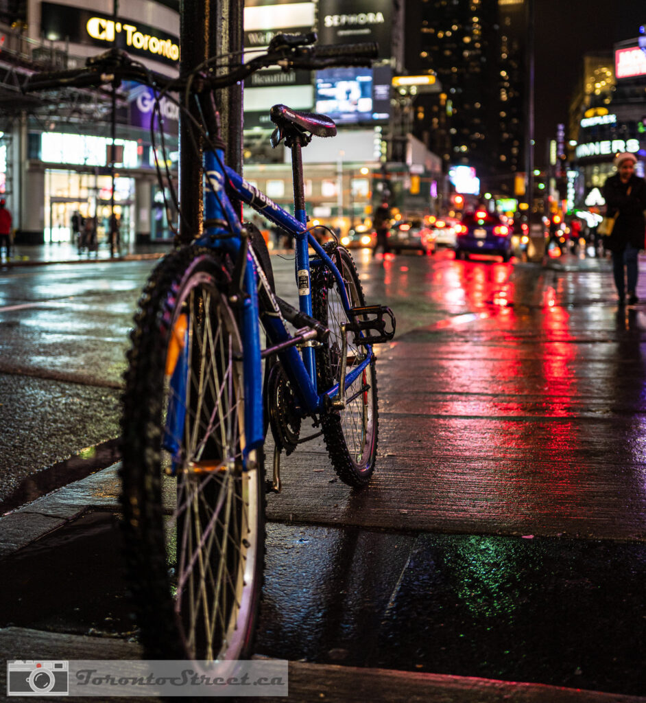 Blue bicycle on rainy Yonge Street at night, red and green neon reflections in wet pavement, colourful Toronto street photography, Fuji X100V