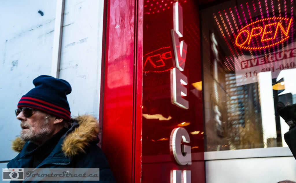 Man in winter coat passing bright red Five Guys storefront with neon Open sign, colourful Toronto street photography, Leica Q2