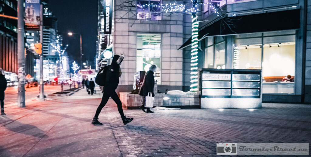 Woman striding on Toronto street at night in winter, Christmas lights and warm storefront glow, shot with Leica Q2