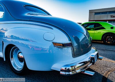 Pale silver rear fender and chrome bumper, a green muscle car and tire shop behind, Leica Q3
