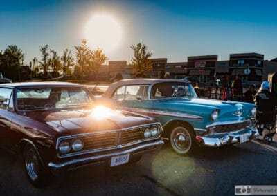 Turquoise-and-white classic beside a maroon coupe as the sun flares over a suburban plaza, people in camp chairs, Leica Q3