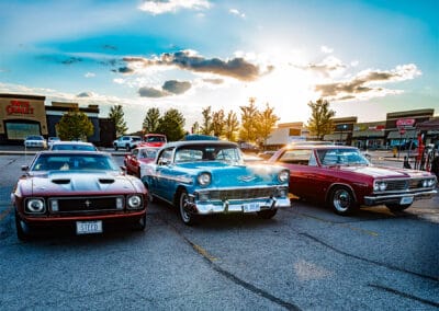 Three classic cars in a row at golden hour, a Mustang, a turquoise drop-top and a maroon coupe, sunburst behind, Leica Q3