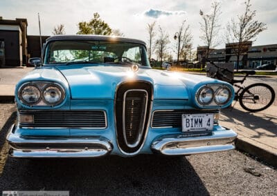 The blue coupe under a dramatic evening sky, chrome grille glowing, a bicycle parked behind, Leica Q3