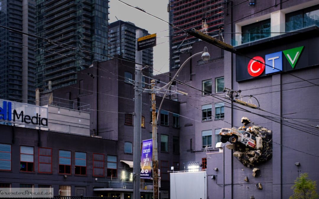 Stock-car sculpture bursting out of the CTV building wall above Queen. What a colourful city street photography opportunity for a Fuji X100V