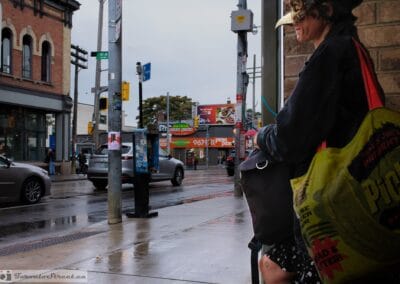 Woman in a bird-mask headpiece at a Danforth corner in the rain, bright bag and jacket, toronto street photography interesting people, Fuji X100V