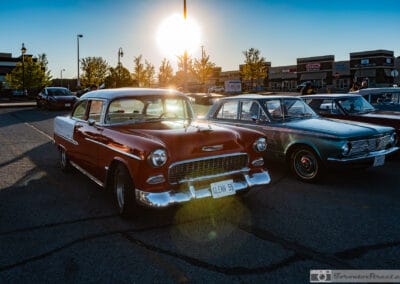 Red two-tone hardtop and a compact sedan beside it as the sun bursts over a suburban plaza, Leica Q3