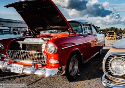 Owner standing by his red hardtop with the hood up under a cloudy sky, a yellow muscle car behind, Leica Q3