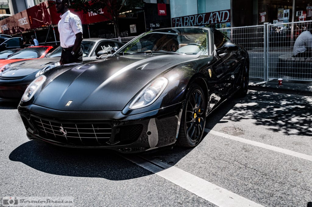 Ferrari 599 GTO--1001040 Two-tone black supercar at the curb with an event marshal standing by, an older red exotic parked behind, Leica Q3