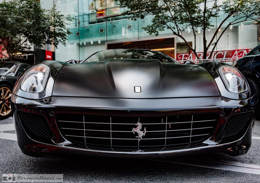 Ferrari 599 GTO--2-3 Black Ferrari 599 GTO head on, twin round headlights and grille badge, storefronts behind, Leica Q3