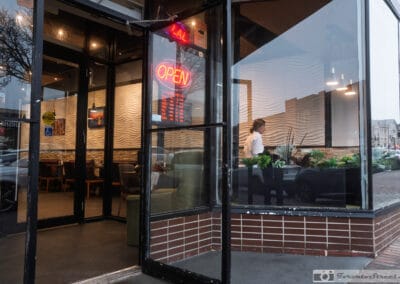 Red OPEN neon on a corner restaurant door at dusk, warm wavy-wood interior glowing through the glass, Leica Q3