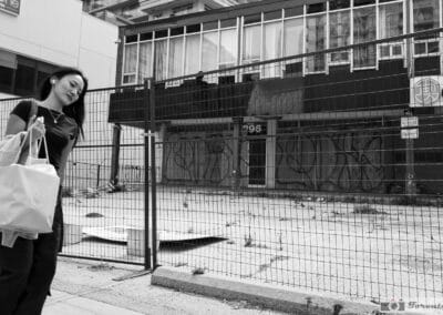 Young woman with earbuds walking past a construction fence downtown, black and white, Leica Q2