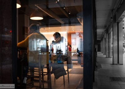 A man in a mustard sweater at a cafe counter seen through the glass, the empty sidewalk colonnade beside him, Leica Q3