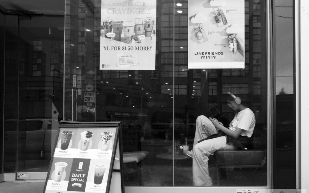 Toronto window reflections over a bubble tea shop, a person in headphones on the bench checking a phone, Leica Q3