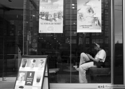 Toronto window reflections over a bubble tea shop, a person in headphones on the bench checking a phone, Leica Q3