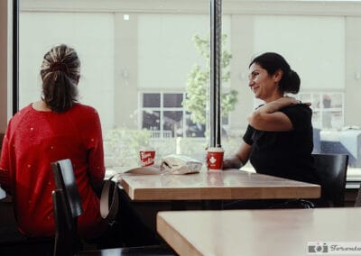 Two women at a Tim Hortons table, one smiling warmly, red cups between them, soft window light, Leica Q3