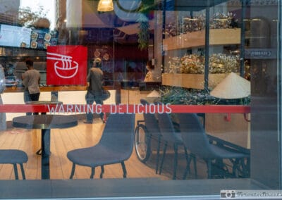 Warning Delicious in red across a ramen shop window, grey chairs and diners inside downtown, Leica Q2