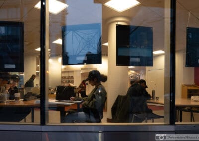 Rows of people at library study tables behind glass, a hydro tower floating across the panes at dusk, Leica Q2