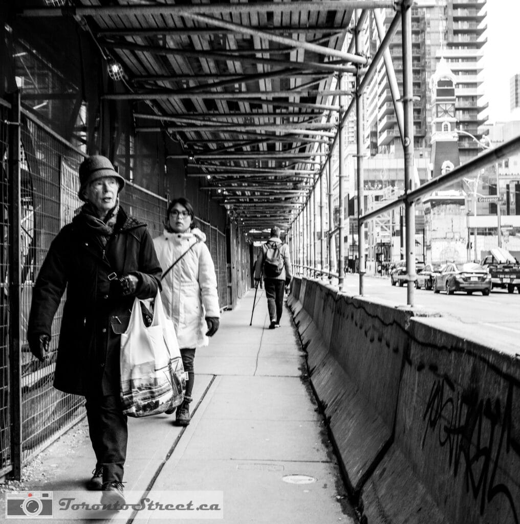 Street Nov 16 2019-1000820 Woman in hat walking through covered construction walkway, black and white street photography Toronto, Leica Q2