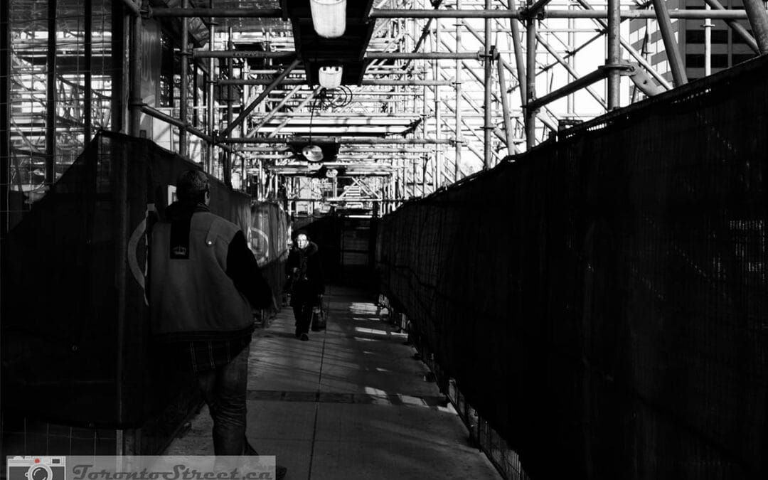 Lone figure lit by single work lamp in dark Toronto construction corridor, black and white street photography, Leica Q2