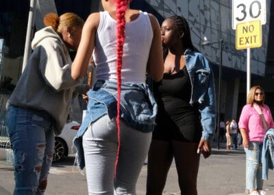 Three young women on a downtown street, one with a long bright pink braided ponytail, glass facade behind, Fuji X100V