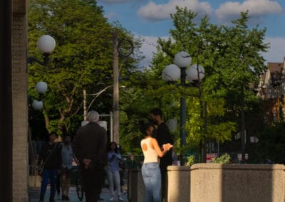 Couple talking in warm golden evening light near a downtown public space, green trees behind them, Fuji X100V