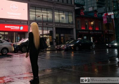 Woman in all-black with long straight blonde hair on a wet downtown sidewalk at night, Shoppers Drug Mart neon behind her, Leica Q2