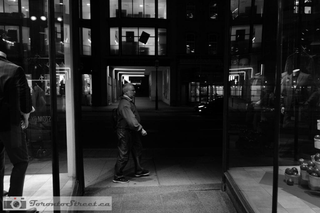 TorontoStreet-L1000544 Man in doorway between suit shops at night, geometric corridor of lit arches behind him, Leica Q2