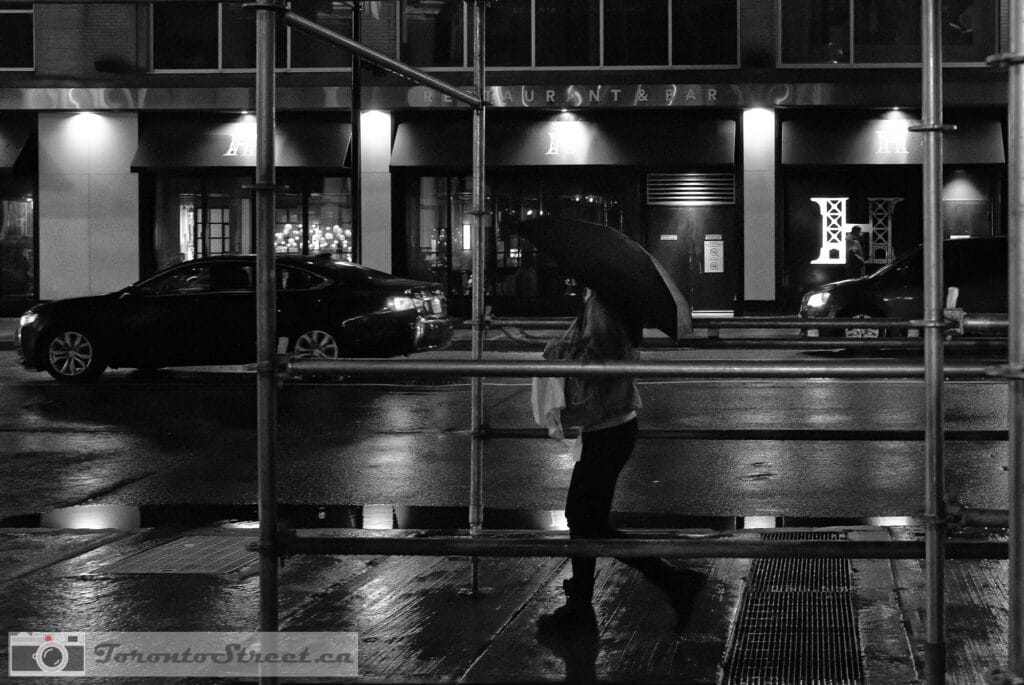 TorontoStreet-L1000655-Edit-Edit Woman with umbrella on rain-slicked Toronto street at night, shot through scaffolding bars Leica Q2