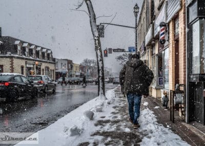January snowstorm in Richmond Hill, a man in a winter jacket walking past shops with a barber pole, Leica Q2