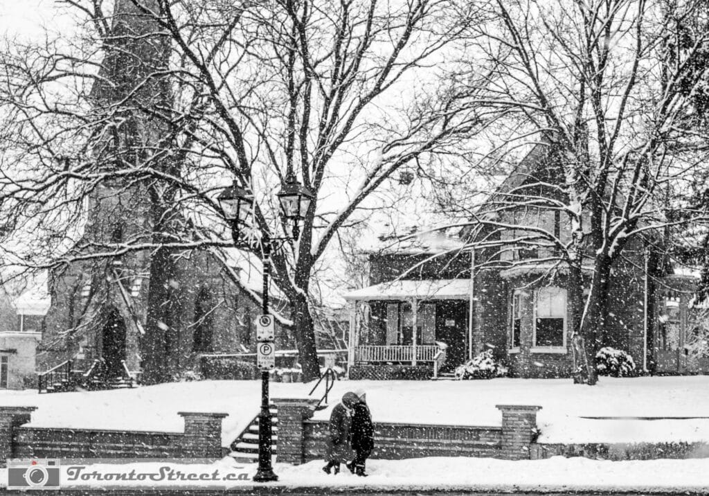 TorontoStreet-L1010413 Two people under umbrella in heavy snowfall outside Toronto church, bare winter trees, Leica Q2