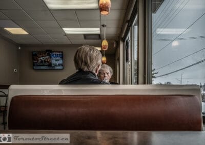 Two elderly friends in close conversation in a Tim Hortons booth, shot through the window from the sidewalk, Leica Q2