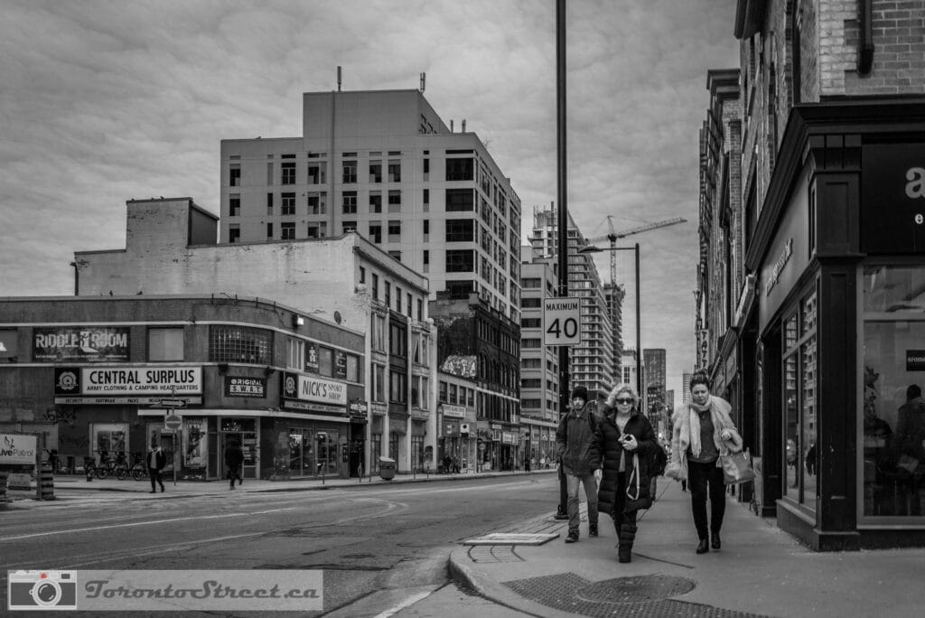 TorontoStreet-L1010432 Three pedestrians on Toronto street with dramatic clouds, Central Surplus store in background, black and white, Leica Q2