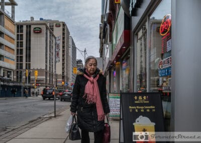 Older woman in a black coat with a pink scarf walking past the Xing Fu Tang tea shop in Toronto, Leica Q2