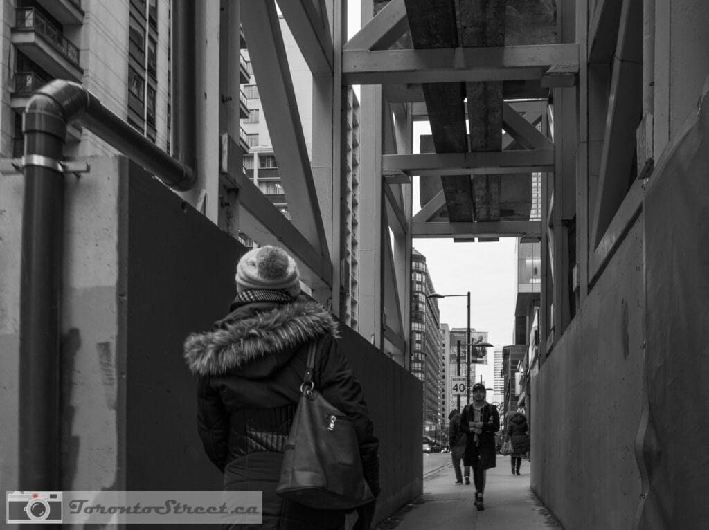 TorontoStreet-L1010455 Woman in winter hat in narrow construction passage with massive steel beams overhead, black and white Toronto, Leica Q2