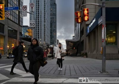 Two women crossing at a downtown intersection at dusk, orange pedestrian signal and a Chanel billboard above, Leica Q2