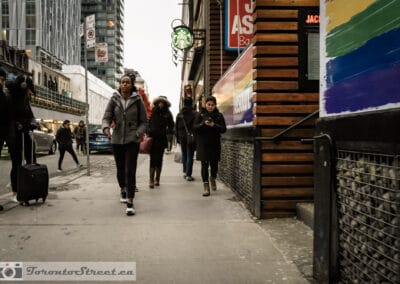 Stream of pedestrians on Yonge Street near a Starbucks, a woman with a rolling suitcase in the foreground, Leica Q2