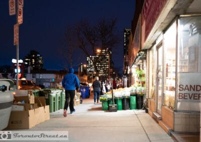 Man in a blue jacket jogging at blue hour past a flower shop, city lights in the background, Leica Q2