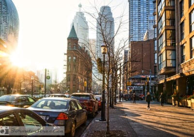 Toronto sunny day street photography, Gooderham Flatiron Building in golden winter light, bare trees and parked cars along the block, Leica Q2
