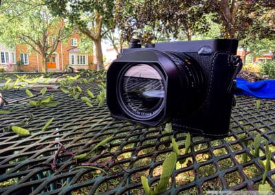 Leica Q3 front view, red Leica dot taped over for security, 28mm lens hood, brick house behind, Galaxy Z Fold7