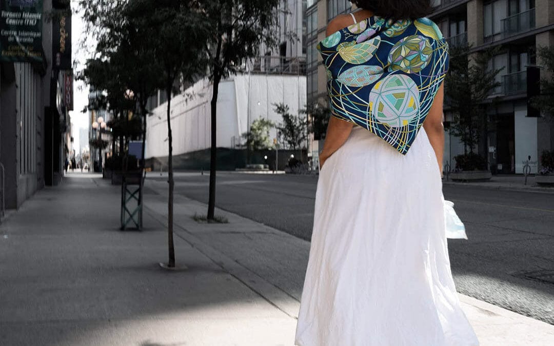 Woman in a long white skirt and a blue and green patterned shawl walking away down an empty downtown sidewalk in late golden light, Toronto, Leica Q2