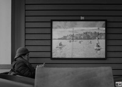 Black and white, a person seated alone at a window table on their phone, a Canadian canoe scene on the wall behind, Leica Q3