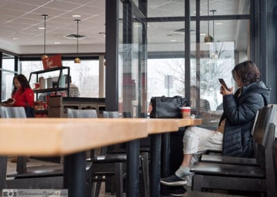 Woman in a puffer jacket on her phone at a window seat, snowy parking lot outside, another woman reading at the counter, Leica Q3