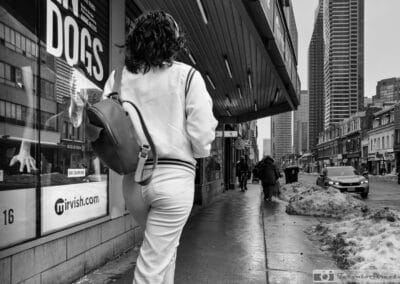 Black and white, a woman in a white tracksuit and backpack passing a theatre window in the slush, Leica Q3