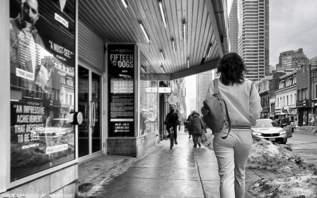 Toronto winter street photography in black and white, a woman walking under a marquee past a cyclist on snowy Yonge, Leica Q3