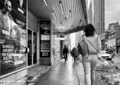 Toronto winter street photography in black and white, a woman walking under a marquee past a cyclist on snowy Yonge, Leica Q3
