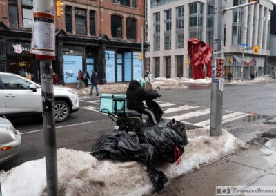 A food-delivery scooter parked by a snowbank piled with garbage bags, a red sculpture across the road, Leica Q3