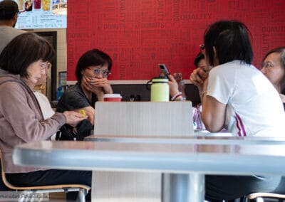 Four women in animated group conversation at a Tim Hortons table, bold red brand wall behind them, Leica Q3