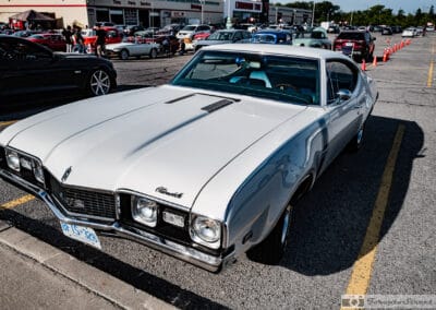 White hardtop coupe at the head of a row of show cars, traffic cones and a tire shop behind, Leica Q3